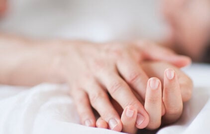 Man sieht die Hände einer Frau und eines Mannes, die zärtlich verschränkt sind. Die Hände liegen auf einem weißen Bettlaken, das Bild hat eine erotische Atmosphäre.