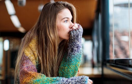 Eine junge Frau sieht gedankenverloren beim Fenster hinaus. Sie hat brünettes, langes Haar, strahlend blaue Augen und trägt einen bunten Strickpullover.